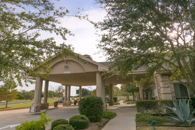 Entrance area of Legend Oaks Healthcare and Rehabilitation - North Austin featuring a covered drop-off zone with columns, surrounded by landscaped bushes and trees under a partly cloudy sky.