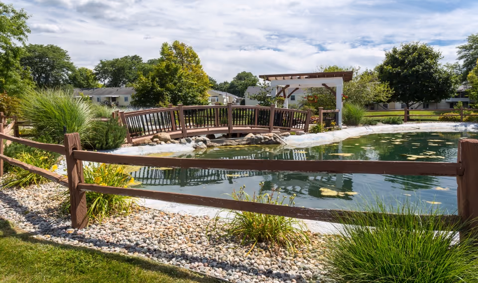 A landscaped outdoor pond with a wooden footbridge, pergola, split-rail fence, and surrounding trees and shrubs on a sunny day.
