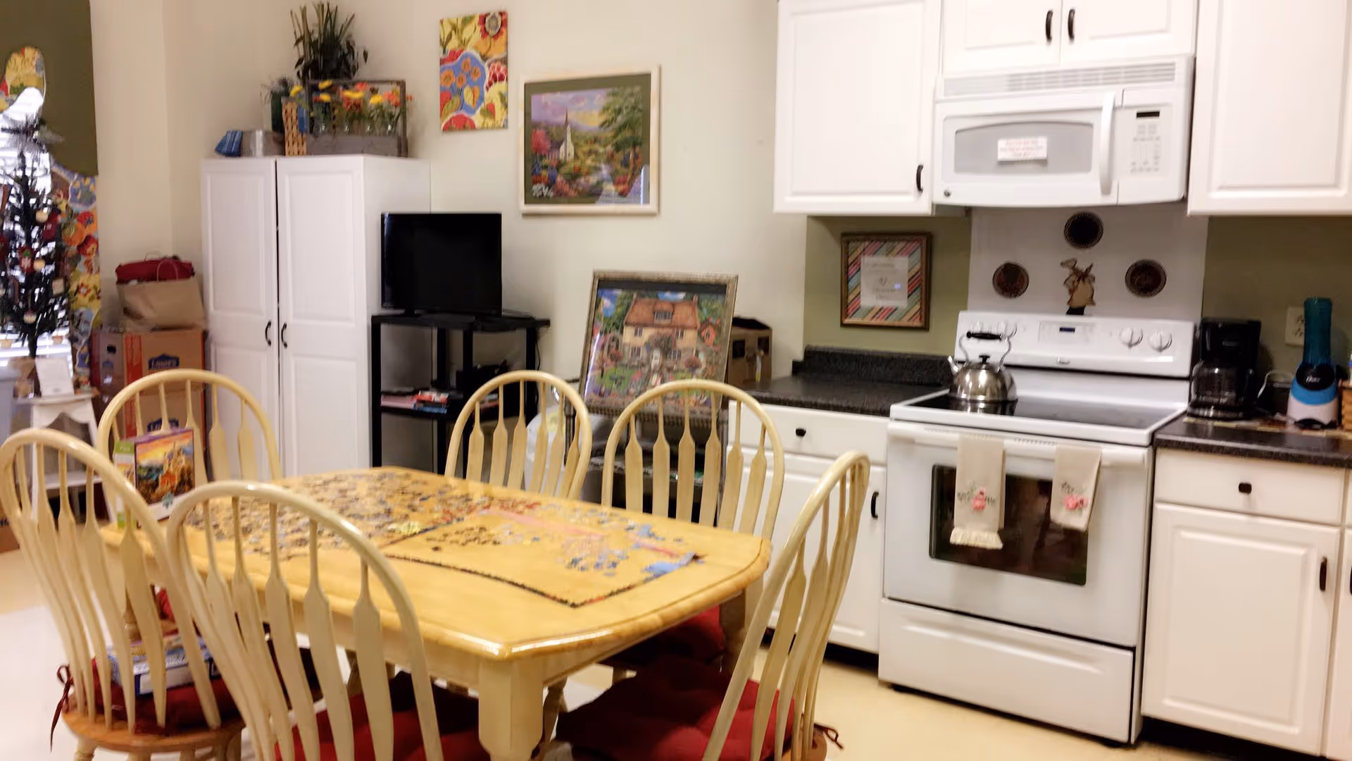 A cozy kitchen and dining area with a wooden table and six chairs. The table has a puzzle in progress on it. The kitchen features white cabinets, a white stove with a kettle on it, a microwave, a coffee maker, and a blender. There are colorful paintings and decorations on the walls and a small TV on a black stand near a white cabinet.
