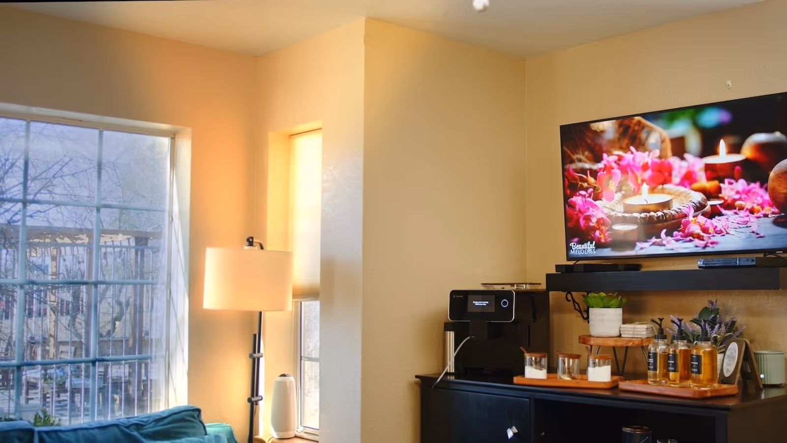 A cozy corner of a room with a large window and a smaller window letting in natural light. There is a floor lamp between the windows. A black cabinet holds a coffee machine, various jars, bottles, and small potted plants. Above the cabinet, a flat-screen TV displays an image of lit candles surrounded by pink flowers.