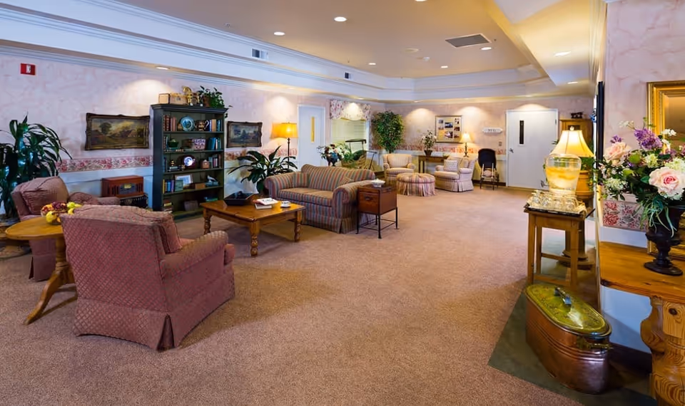 A cozy and well-lit living room area in a senior living facility with multiple upholstered armchairs and sofas arranged around wooden coffee tables. The room features soft pink walls with floral wallpaper borders, various plants, framed paintings, and decorative lamps. There is a bookshelf filled with books and decorative items, and a water dispenser on a small table near the entrance door.