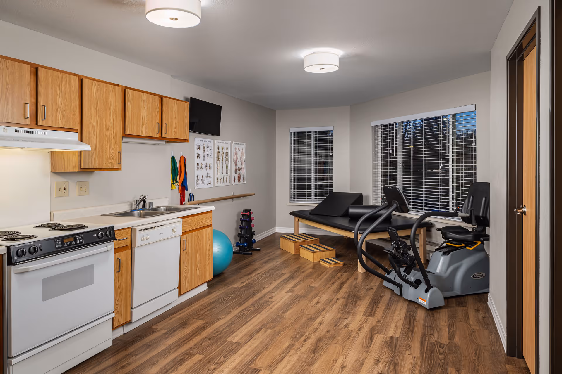 A room with a kitchen area on the left featuring wooden cabinets, a stove, dishwasher, and sink. The right side of the room has exercise equipment including a recumbent bike, a therapy table, exercise balls, resistance bands, and anatomical charts on the wall. The room has wood flooring, white walls, and two windows with blinds.
