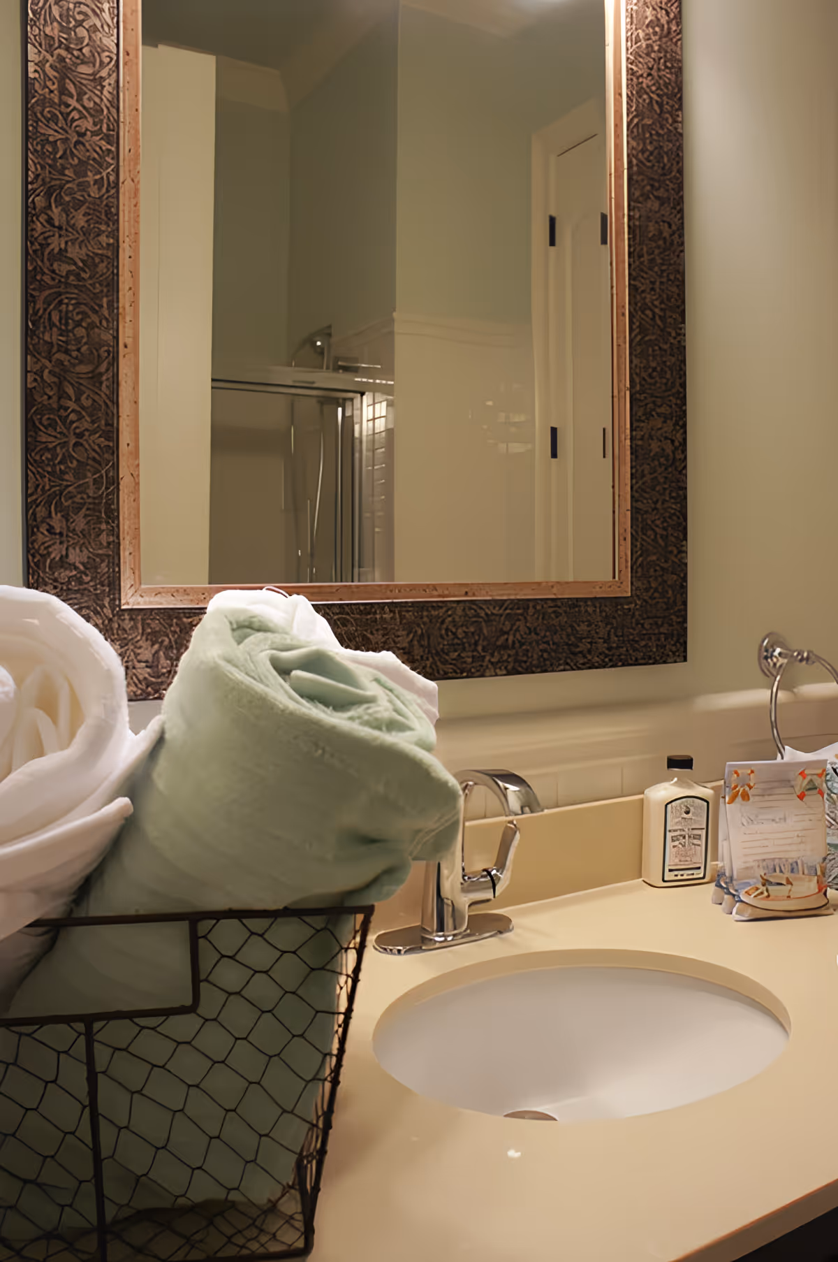 Bathroom vanity with sink, faucet, rolled towels in a wire basket and a framed mirror reflecting a shower.