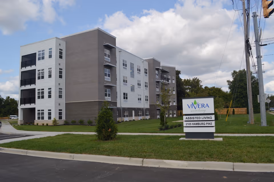 Exterior view of a modern four-story assisted living building with balconies and multiple windows, surrounded by green grass and a paved driveway. A sign in front reads 'Vivera Senior Living, Assisted Living, 2105 Hamburg Pike'. The sky is partly cloudy.