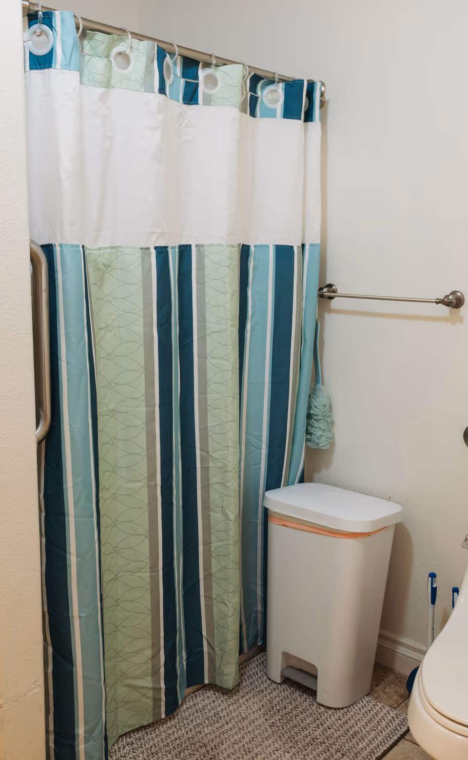 A bathroom corner featuring a shower with a striped shower curtain in shades of green, blue, and white. Next to the shower is a white trash bin with a lid, a towel rack without a towel, and a light green bath sponge hanging on the wall. The floor has a textured bath mat.