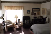A cozy bedroom with a large bed on the right, a black recliner chair, a small table with a lamp, and a cushioned armchair near a window with sheer curtains and a valance. The walls are decorated with framed pictures and maps.
