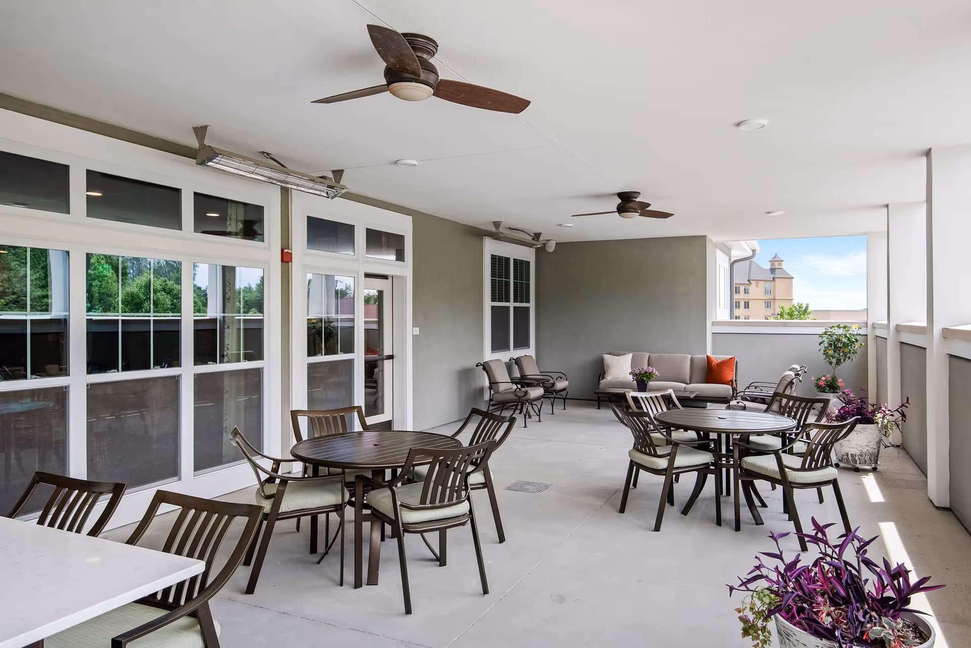 Covered outdoor patio area with several round tables and chairs, ceiling fans, and a seating area with a sofa and armchairs. Potted plants are placed along the edges, and large windows and doors lead inside the building.