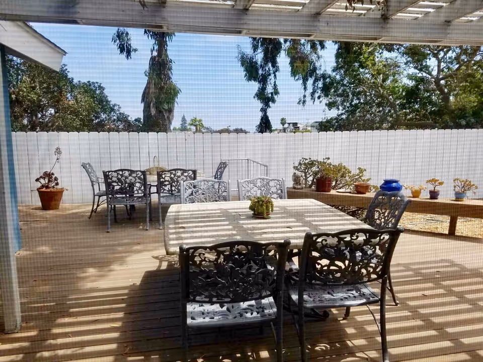Outdoor patio area with metal tables and chairs featuring intricate designs. Several potted plants are placed on the tables and along a wooden bench against a white fence. Trees and greenery are visible beyond the fence under a clear sky.