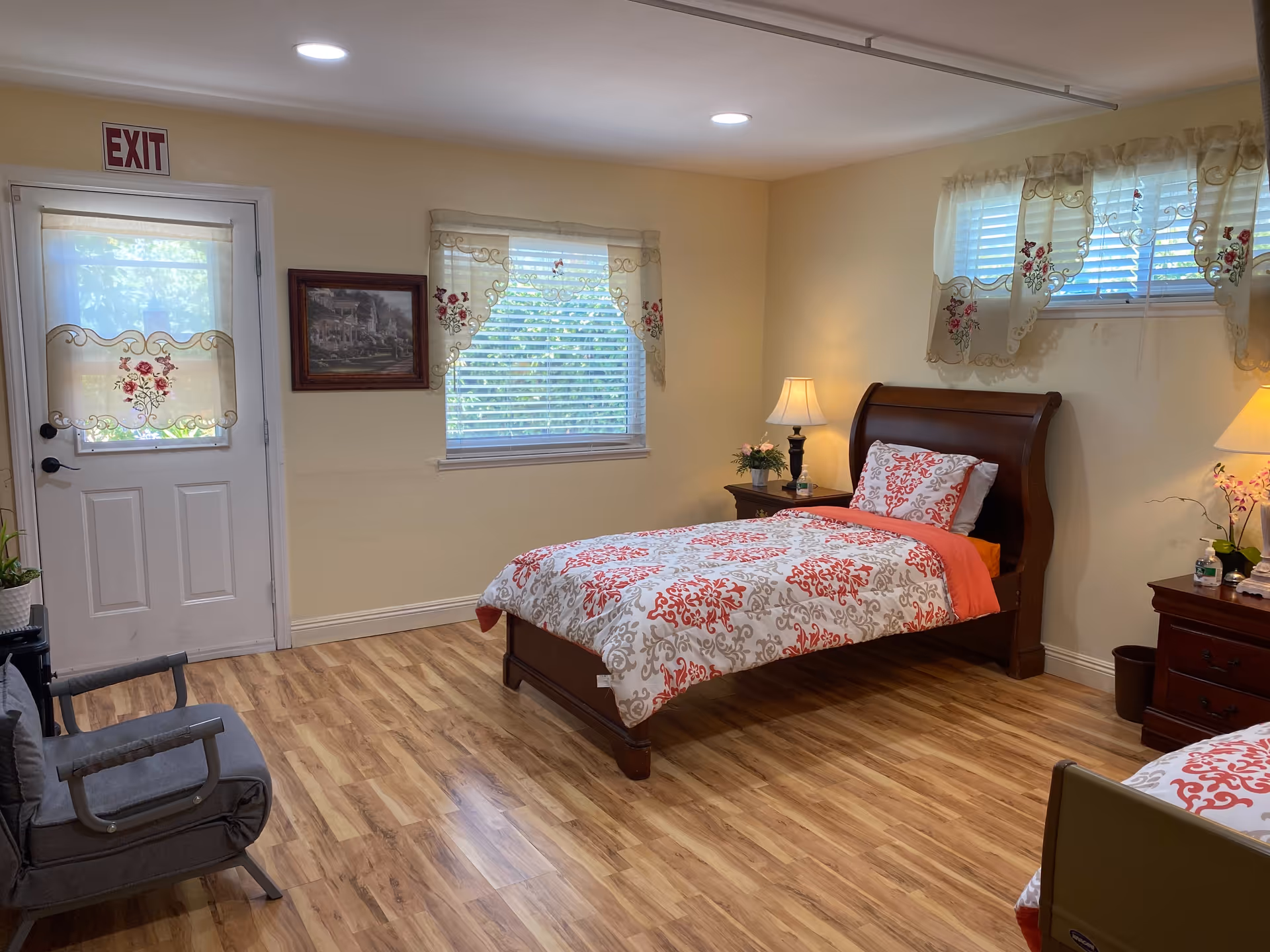 A cozy bedroom with two single beds featuring white and red patterned bedding. The room has wooden flooring, two windows with decorative curtains, a door with an exit sign above it, a gray armchair, two bedside tables with lamps, and some small plants and flowers.