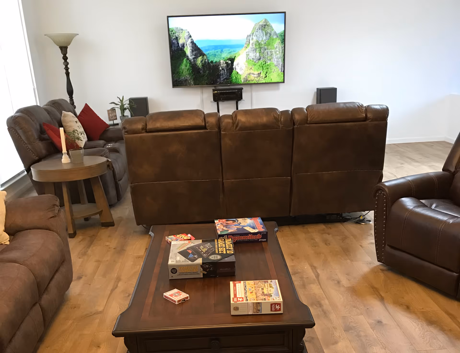 A cozy living room with brown leather recliners and sofas arranged around a wooden coffee table. The coffee table has several board games and a deck of cards on it. A flat-screen TV mounted on the wall displays a scenic image of green mountains. There is a tall floor lamp and a small side table with a plant and decorative items next to one of the sofas. The room has wooden flooring and white walls.