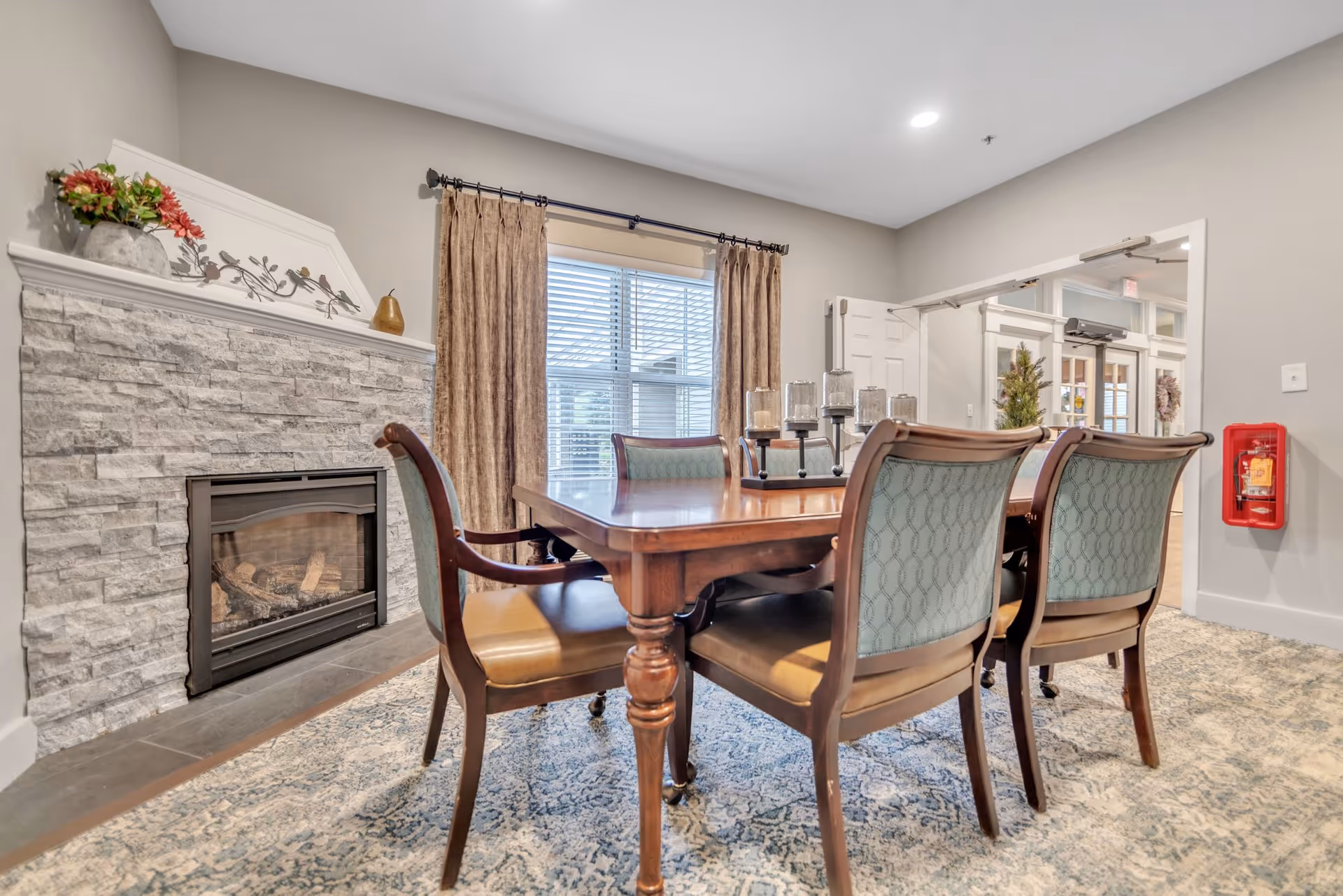 Wood dining table with upholstered chairs in a cozy common room featuring a stone fireplace and window.