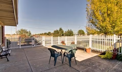 Outdoor patio with a plastic table and chairs, benches, potted plants and a white picket fence with trees beyond.