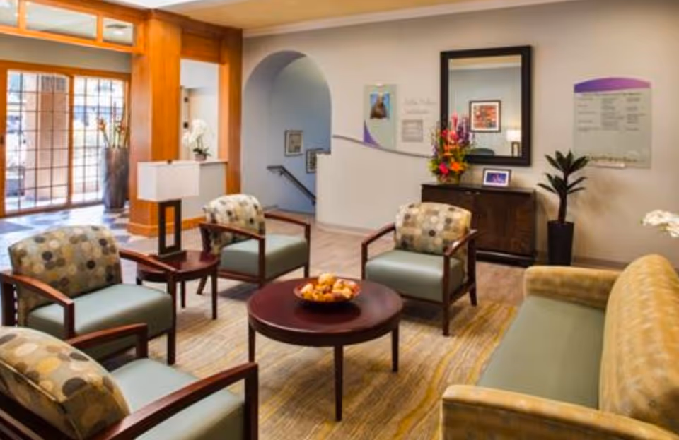 A bright senior living lounge with multiple upholstered armchairs and a sofa arranged around a round coffee table, near a console table and mirror.