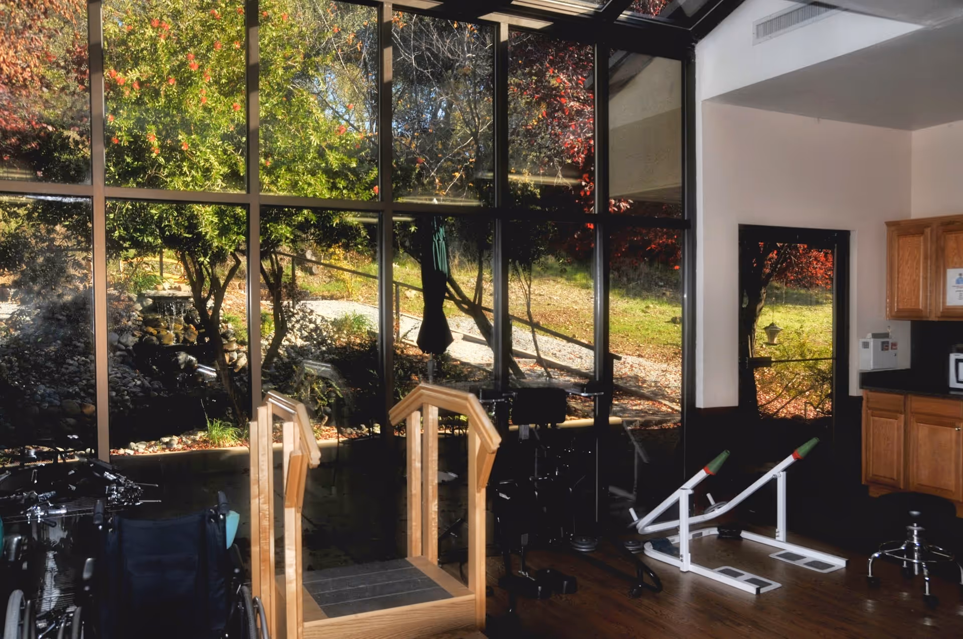 Interior view of a rehabilitation or therapy room with large floor-to-ceiling windows showing an outdoor garden area. The room contains exercise equipment including parallel bars for walking therapy, a wheelchair, and other physical therapy tools. Wooden cabinets and a microwave are visible on the right side.