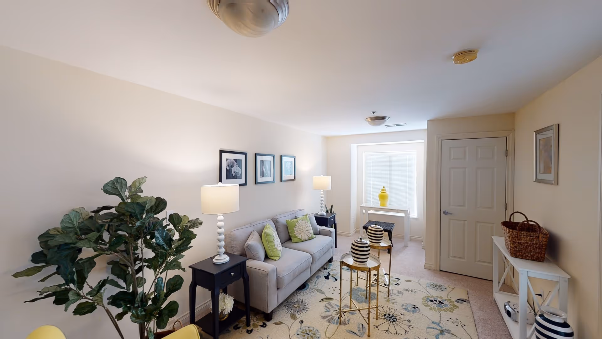 Bright furnished living room with a sofa, side tables and lamps, decorative rug, a large potted plant, and a windowed alcove.
