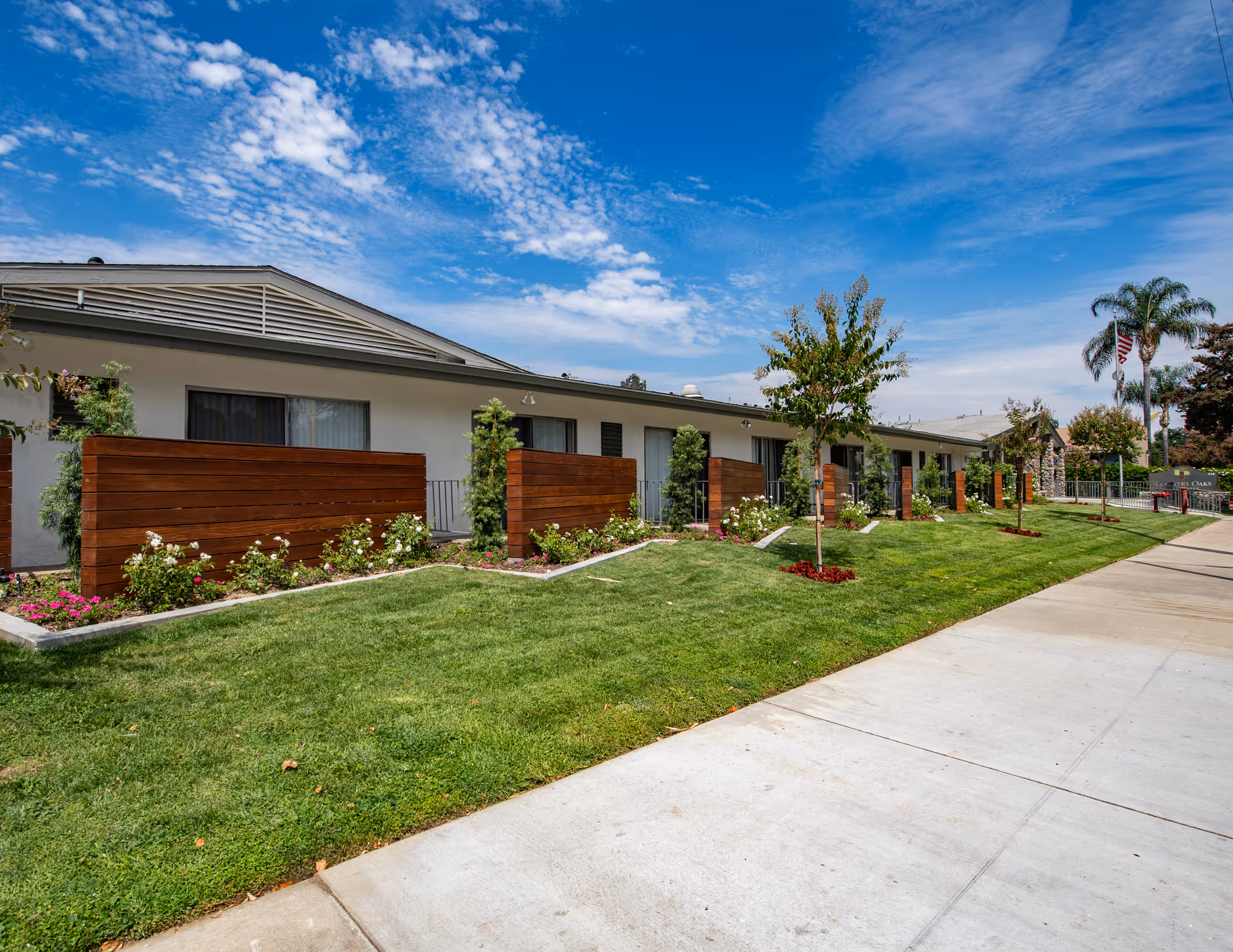 Exterior view of a single-story building with a row of private patios separated by wooden fences. The building is surrounded by a well-maintained lawn, small trees, and flower beds under a partly cloudy blue sky.