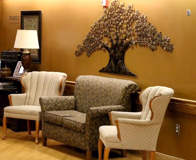 A cozy seating area with two beige upholstered armchairs and a patterned loveseat against a warm brown wall. Above the loveseat is a decorative metal tree wall art. To the left, there is a table with a lamp, framed pictures, and a small shelf. The wall has the word 'FAMILY' and a quote written on it.