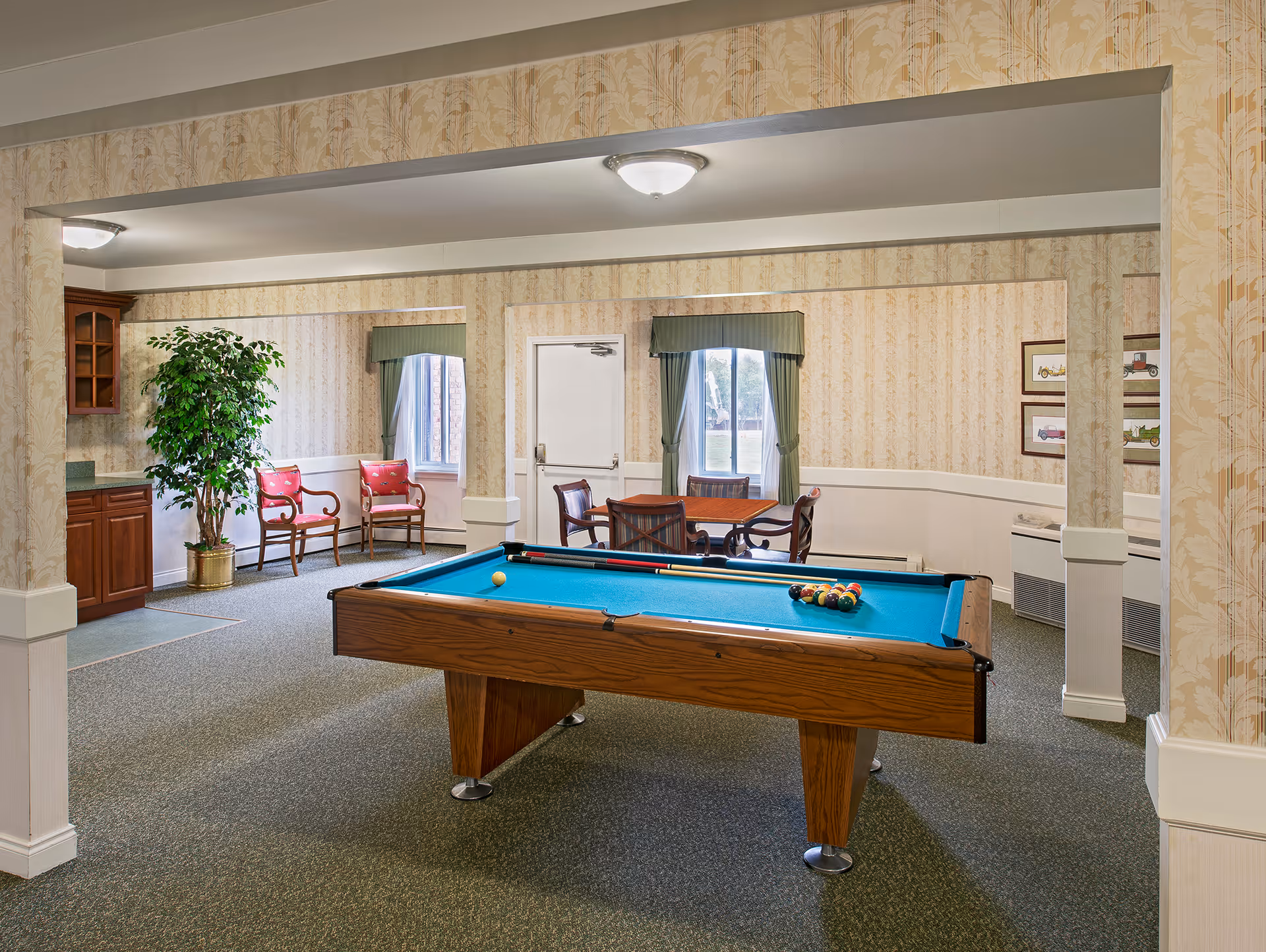A bright communal game room featuring a pool table in the center, chairs and a small table by windows, and patterned wallpaper.