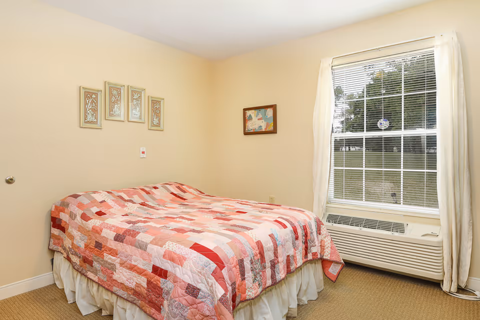 A bedroom with a bed covered in a pink and red patchwork quilt. The room has beige walls with four framed artworks above the bed and one framed artwork on the adjacent wall. There is a large window with white blinds and curtains, and an air conditioning unit below the window. The floor is carpeted in a neutral color.