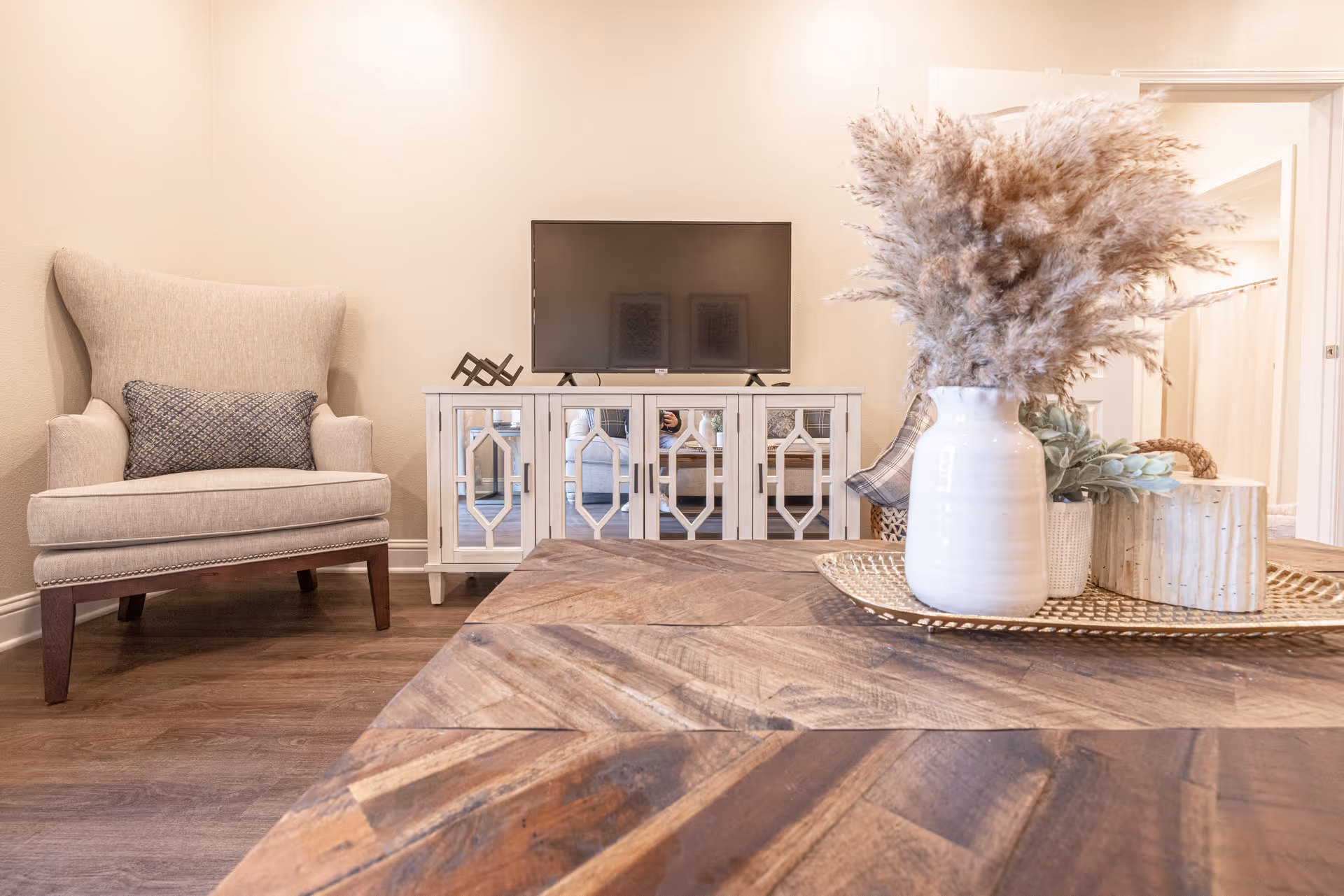 A cozy living room area with a beige armchair with a decorative pillow, a white TV stand with a flat-screen TV on top, and a wooden coffee table in the foreground. On the coffee table, there is a decorative tray holding a white vase with dried pampas grass and other small decorative items. The room has light-colored walls and wood flooring.