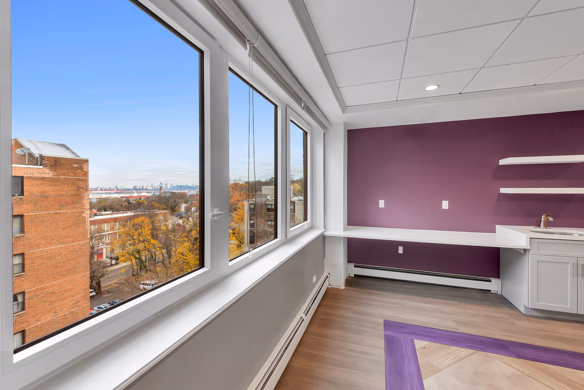 Interior room with large windows showing a city view, a purple accent wall with shelves, and a countertop with a sink.