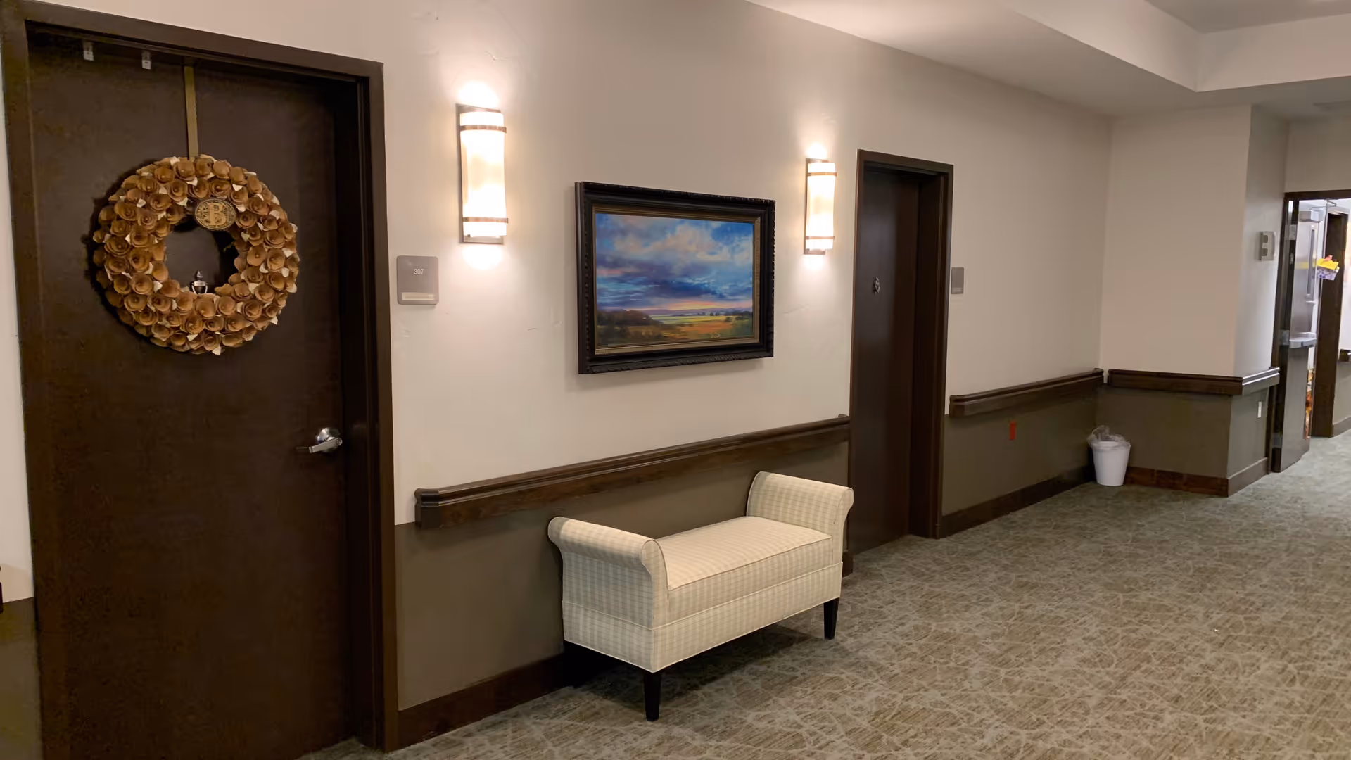 A hallway in an assisted living facility with two dark brown doors, one decorated with a large wreath. Between the doors is a cushioned bench with a checkered pattern, mounted on the wall above it is a framed landscape painting. The walls are painted in two tones, with a chair rail separating the lighter upper section from the darker lower section. The floor is carpeted with a patterned design, and there are wall-mounted lights illuminating the area.