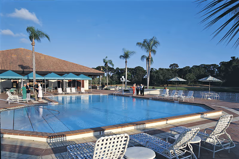 Outdoor swimming pool area at The Barclay at Sarasota with several lounge chairs and tables with umbrellas around the pool. A few people are standing and conversing near the poolside. Palm trees and a building with a tiled roof are visible in the background under a clear blue sky.