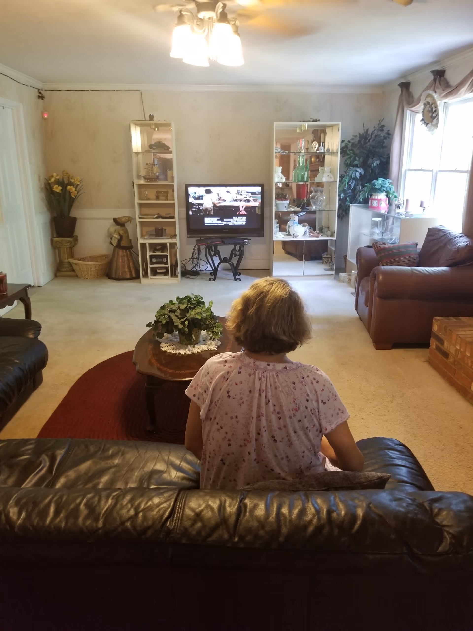 A woman seen from behind sits on a leather couch watching a television in a homey living room with display cabinets and armchairs.
