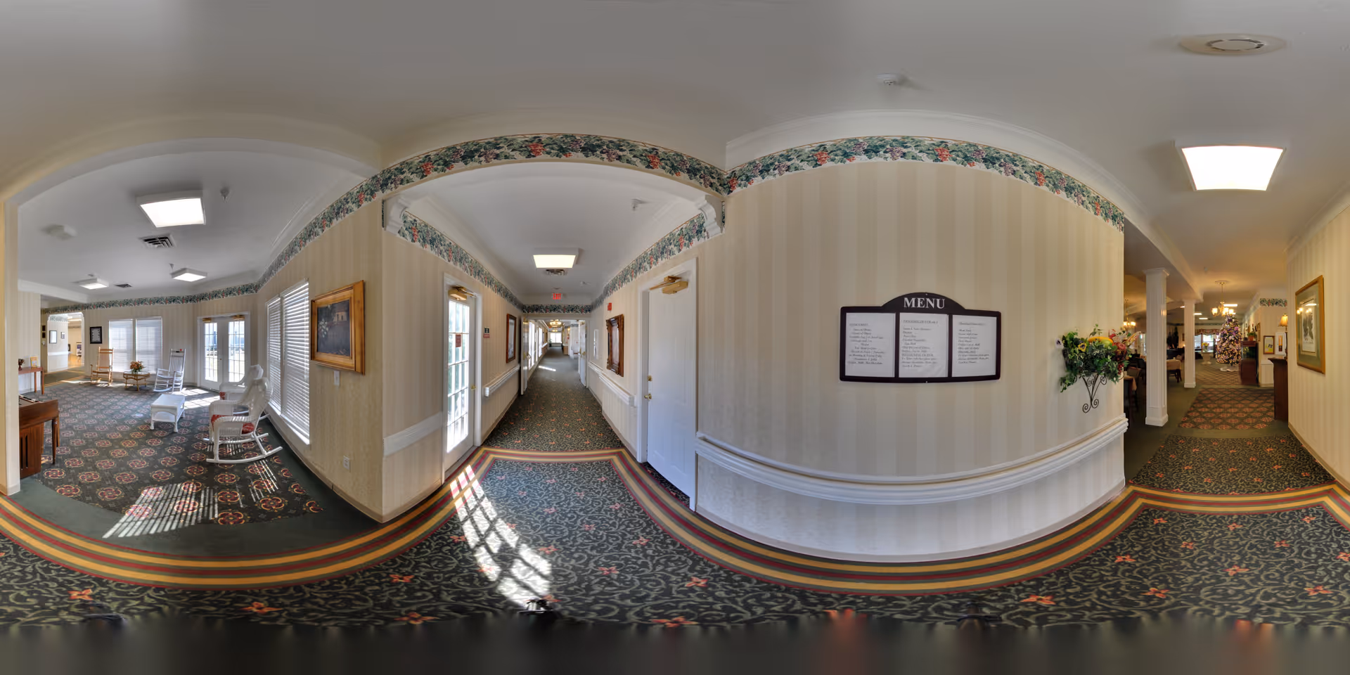 A panoramic view of a senior living facility interior hallway with patterned carpet and floral wallpaper border. On the left side, there is a sitting area with white rocking chairs and windows letting in natural light. The hallway extends forward with doors and framed artwork on the walls. A menu board and a decorative flower arrangement are mounted on the right wall. The area is well-lit with ceiling lights and has a warm, inviting atmosphere.