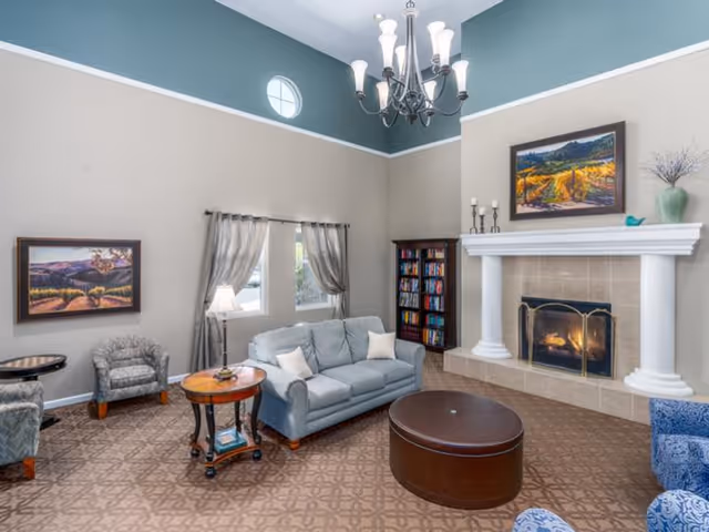 Bright senior living room with high ceilings, chandelier, fireplace, sofa, armchairs, and a round coffee table.