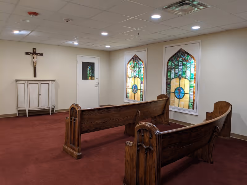 Interior room with red carpet, two wooden pew benches facing a white wall with a crucifix above a small white cabinet. Two stained glass windows are on the adjacent wall, and a white door is between them.