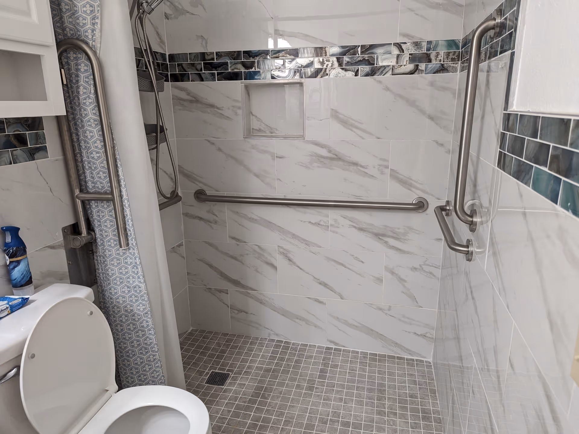 A bathroom with a white toilet, a shower area with marble-patterned tiles, a decorative tile border, and multiple stainless steel grab bars for accessibility. There is a shower curtain with a blue and white pattern and a small built-in shelf in the shower wall.