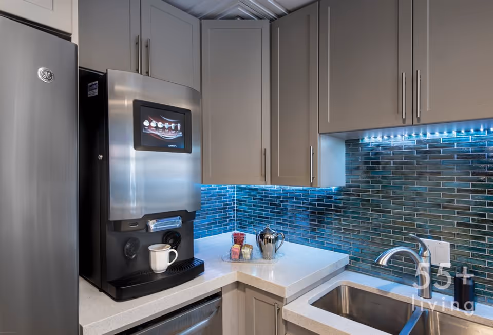 Modern kitchen corner with gray cabinets, a stainless steel refrigerator, a coffee machine with a white mug, a sink with a faucet, and a blue-tiled backsplash illuminated by under-cabinet lighting.
