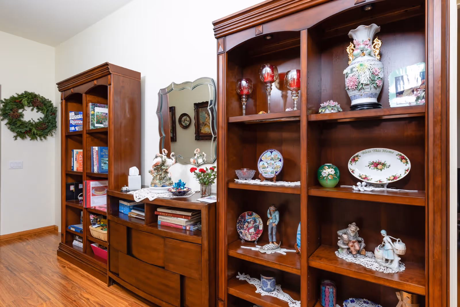 Wooden bookshelves and a dresser with a mirror displaying decorative plates, figurines, books, and vases in a well-lit interior room.