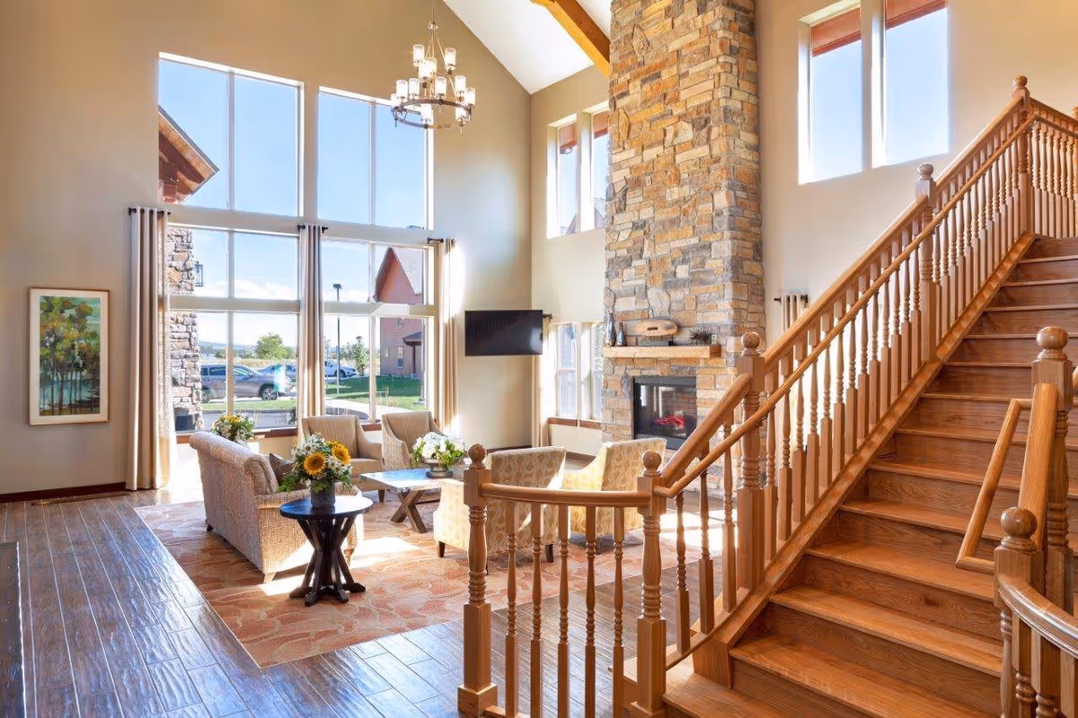A bright and spacious living room area with large floor-to-ceiling windows letting in natural light. The room features a stone fireplace, a wall-mounted TV, comfortable seating including a sofa and armchairs, a wooden side table with a flower arrangement, and a wooden staircase with detailed railings leading to an upper floor.