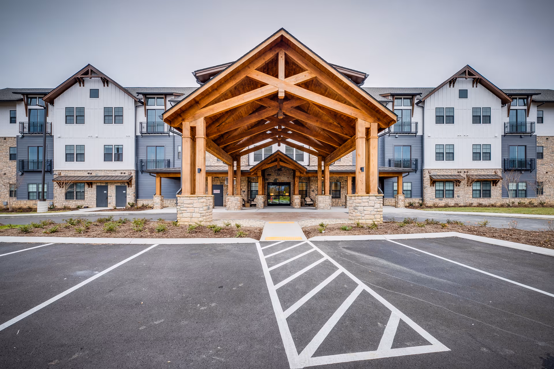 Front exterior view of a large senior living facility building with a prominent wooden covered entrance supported by stone pillars. The building has multiple windows and balconies, with a parking lot in the foreground.