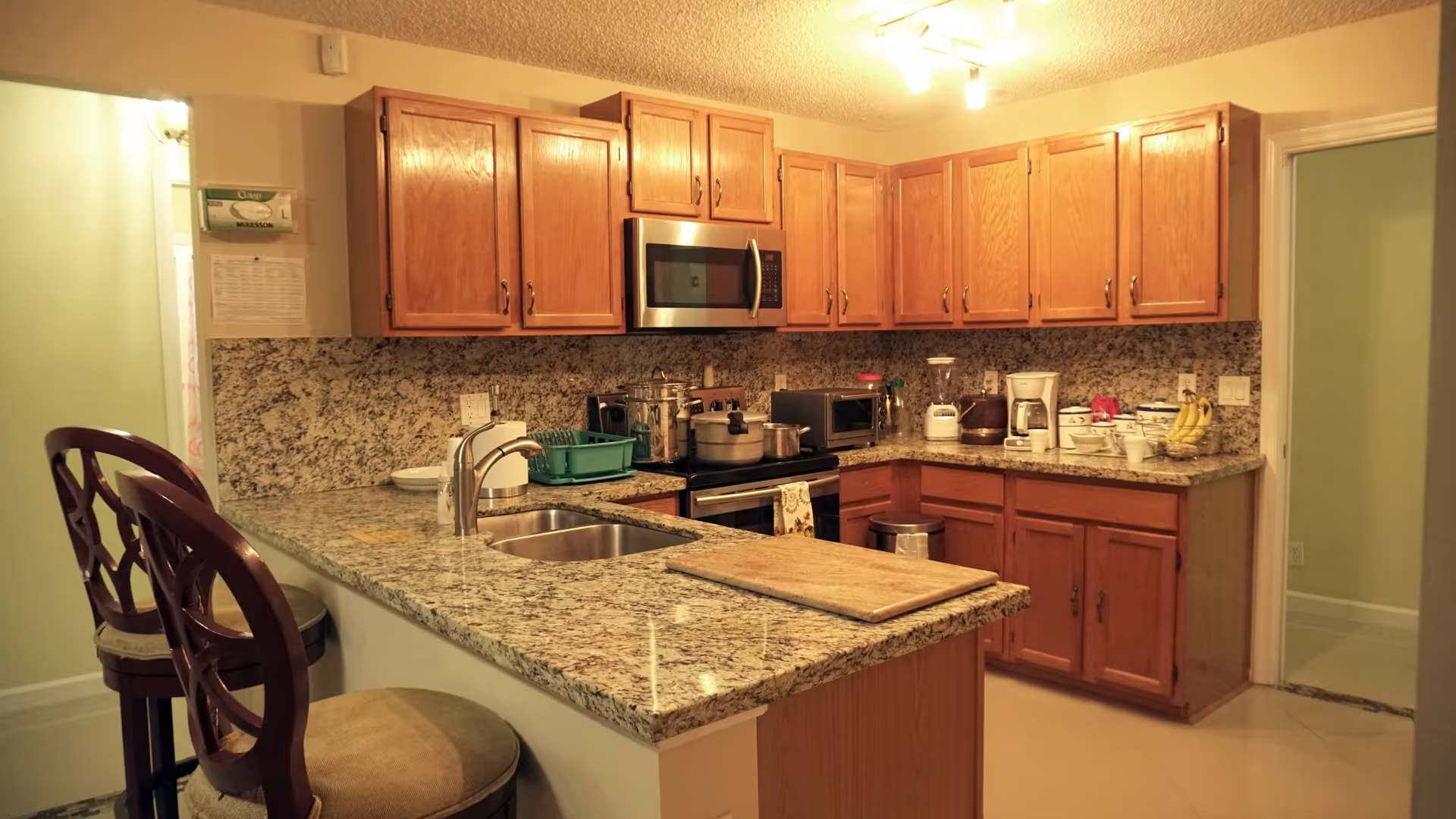 A kitchen with wooden cabinets, granite countertops, and various kitchen appliances including a microwave, stove, blender, coffee maker, and toaster oven. There are two wooden bar stools with cushioned seats at the counter, and a cutting board on the countertop. The kitchen is well-lit with ceiling lights.
