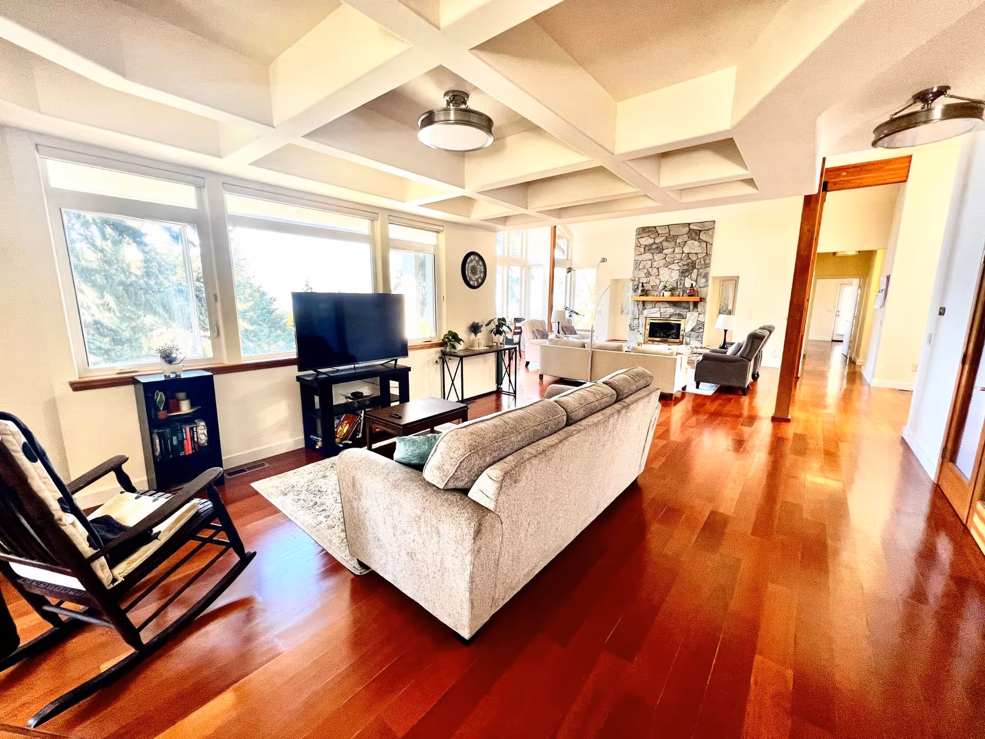 Bright living room with large windows letting in natural light, featuring a beige sofa, a rocking chair, a flat-screen TV on a stand, a coffee table, and a stone fireplace in the background. The room has wooden floors and a coffered ceiling.