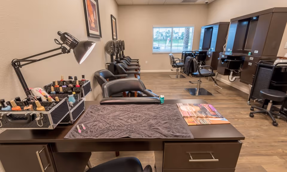 Interior view of a salon area in a senior living facility with manicure and hair styling stations. The foreground shows a manicure table with nail polish bottles, a lamp, and magazines. In the background, there are multiple black salon chairs and hair washing sinks along the wall, with a window letting in natural light.