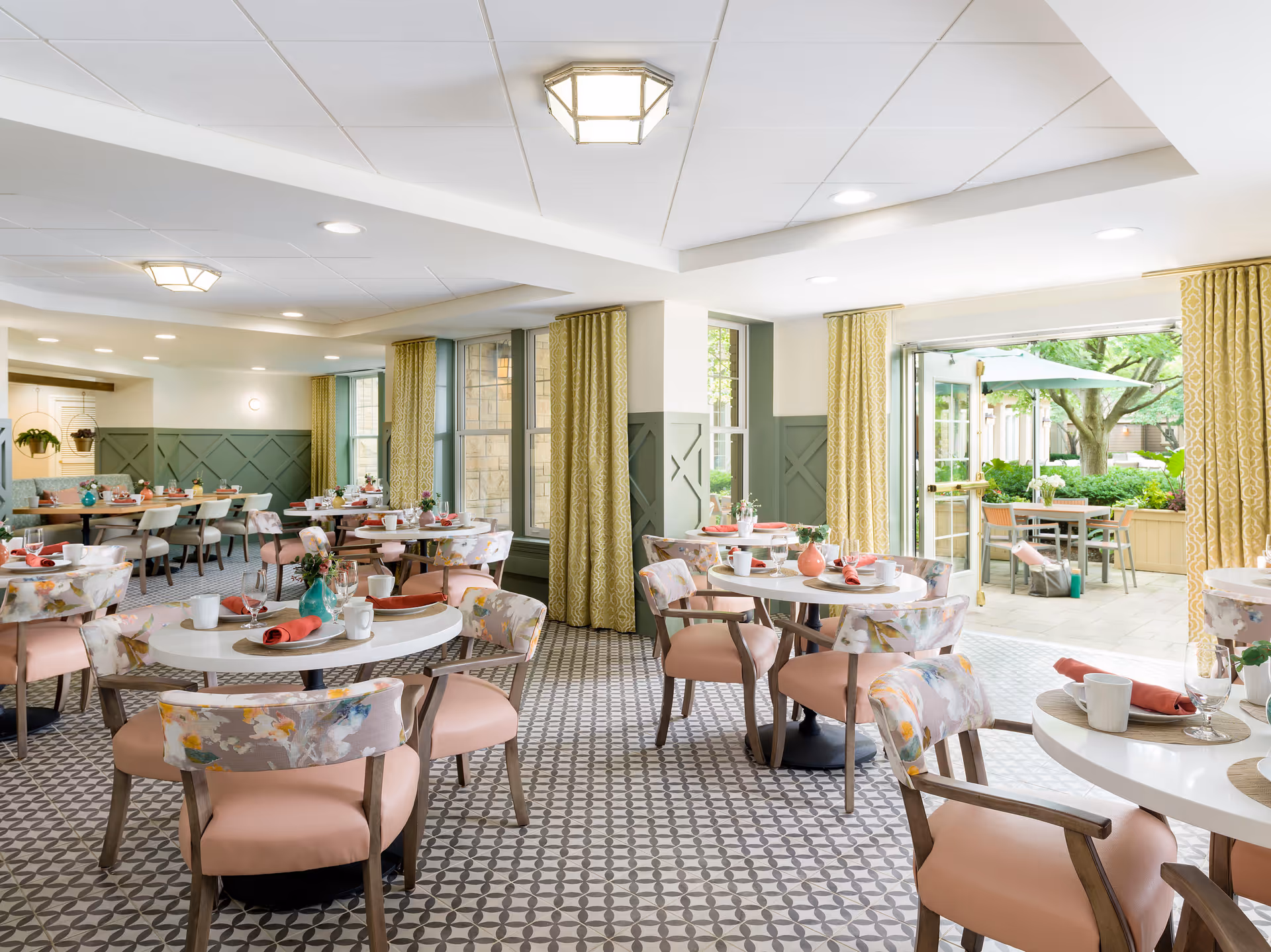 Bright and airy dining room with round tables set with white mugs, plates, and red napkins. The chairs have floral patterned backs and pink cushions. Large windows and glass doors open to an outdoor patio area with tables, chairs, and greenery. The room features patterned tile flooring, green wainscoting, and yellow curtains.