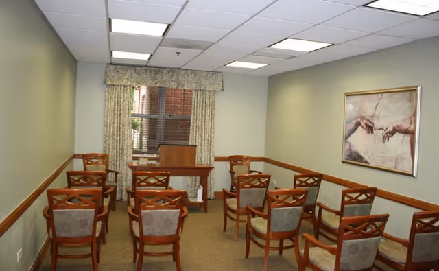Small meeting room with rows of wooden chairs facing a lectern by a window and framed artwork on the wall.