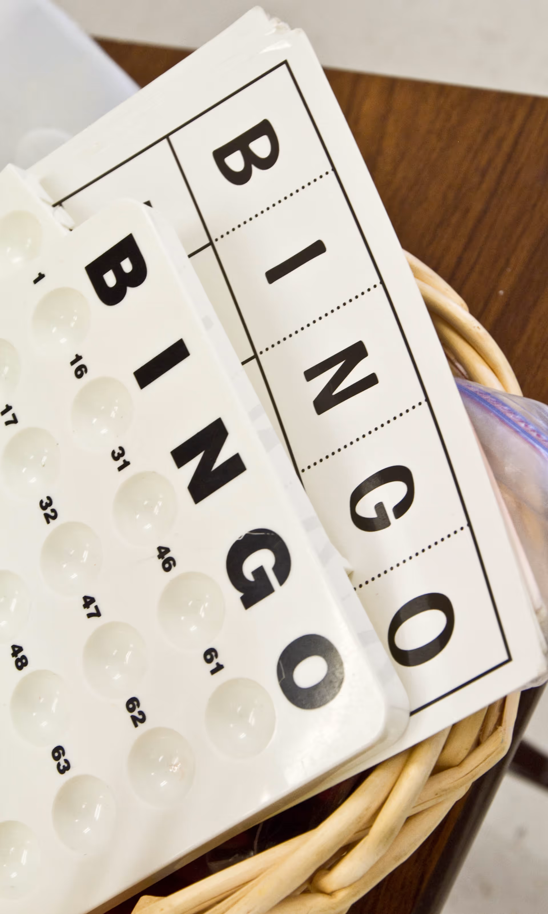 Close-up image of bingo cards and a bingo number holder placed in a wicker basket on a wooden table.