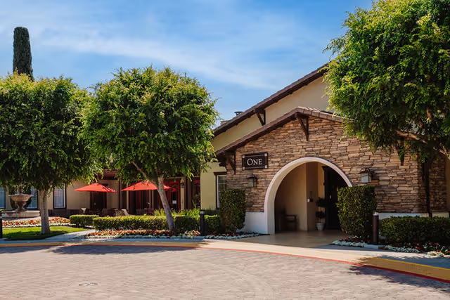 Front entrance of a building with a stone archway labeled 'ONE', landscaped trees, red patio umbrellas and a paved circular driveway.