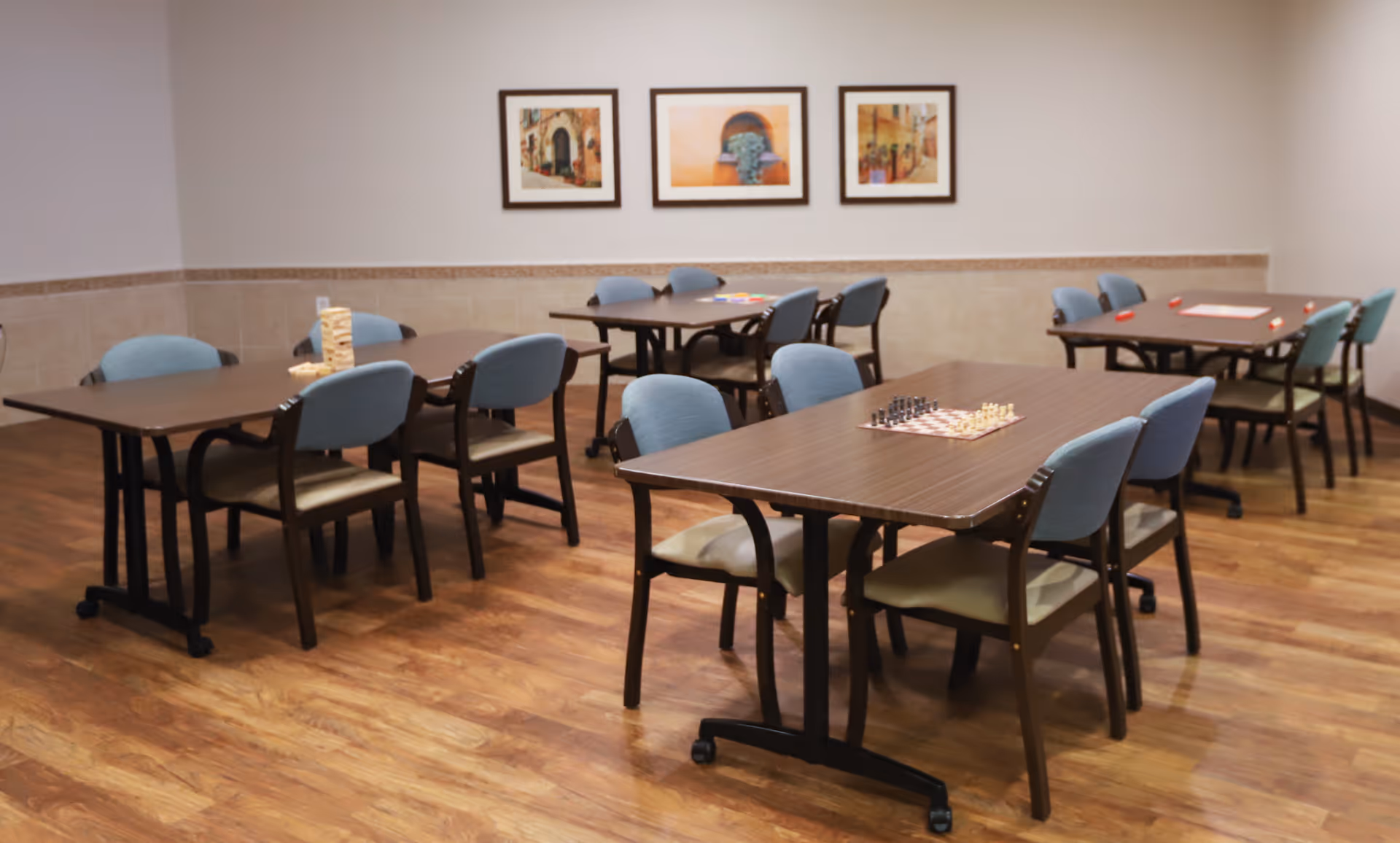 A room with several tables and chairs arranged for group activities. On the tables are board games including chess and Jenga. The room has wooden flooring, beige walls with a decorative border, and three framed pictures hanging on the wall.