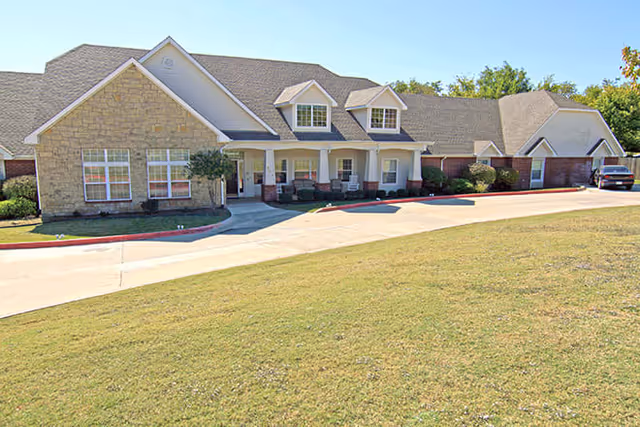 Single-story residential building with a stone and siding exterior, featuring a covered porch with several chairs. The building is surrounded by a well-maintained lawn and a paved driveway leading to a garage on the right side.