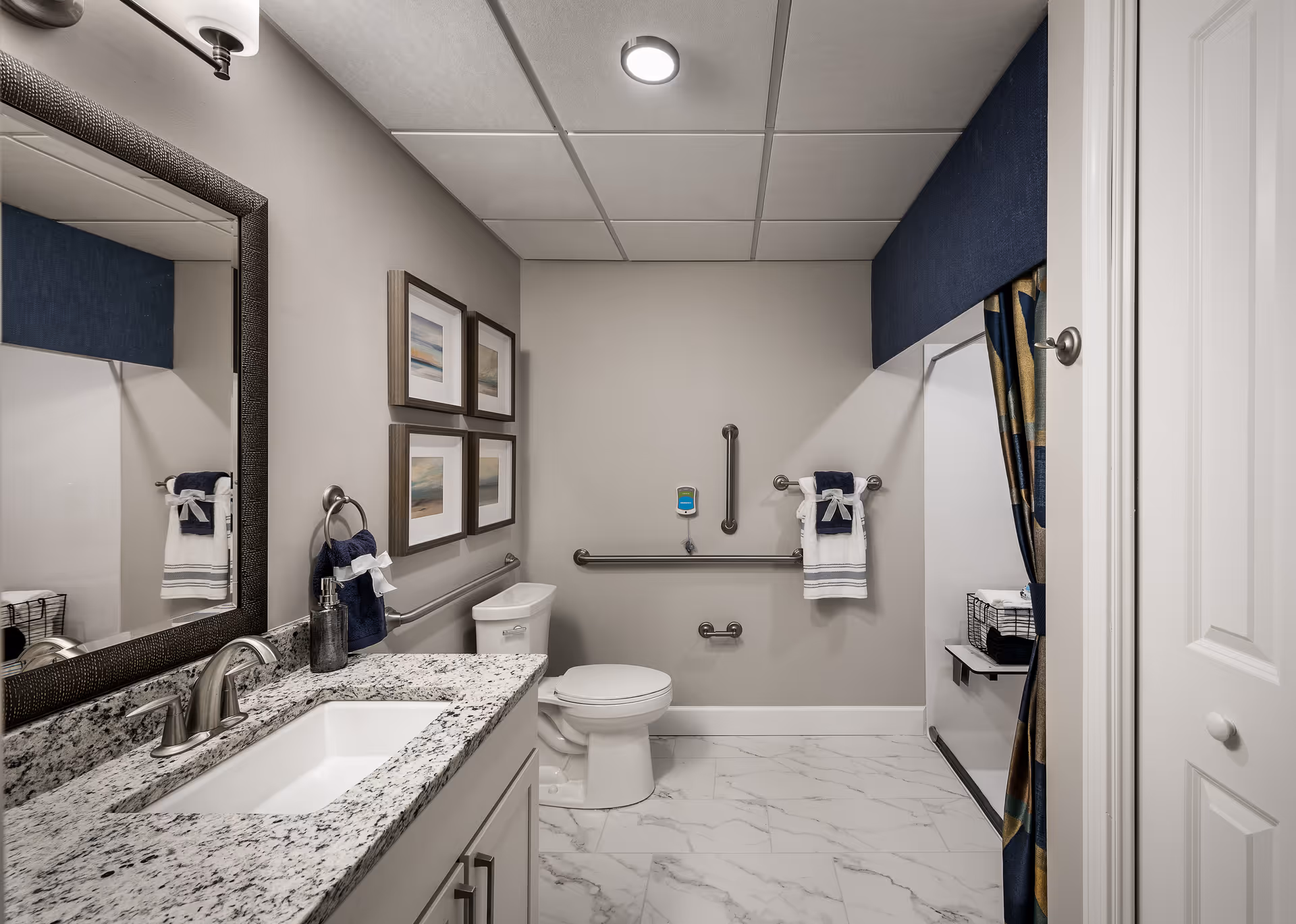 A modern bathroom with a granite countertop sink, a large mirror, and a white toilet. The walls are painted light gray and decorated with four framed pictures. There are stainless steel grab bars around the toilet for accessibility, and a towel rack with neatly folded towels. The floor has white marble tiles, and there is a blue valance above a curtained area on the right side.