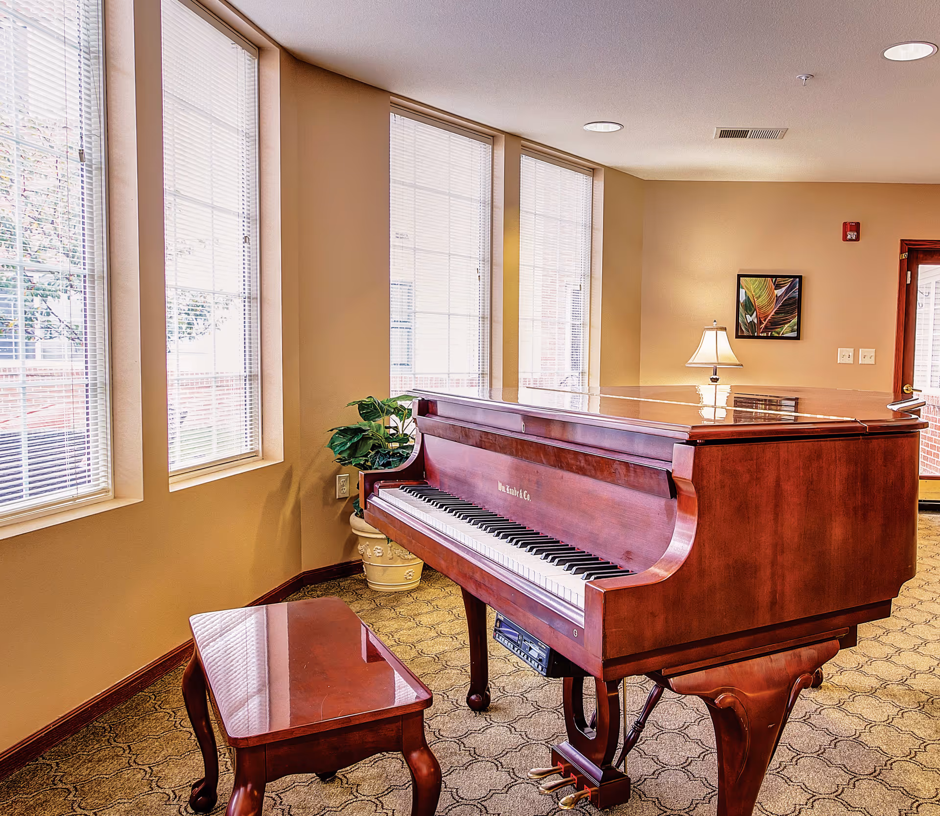 A polished wooden grand piano with a matching bench is placed in a well-lit room with beige walls and large windows covered with blinds. A potted plant sits in the corner near the piano, and a table lamp and framed artwork are visible on the far wall. The room has a patterned carpet and a door leading outside.