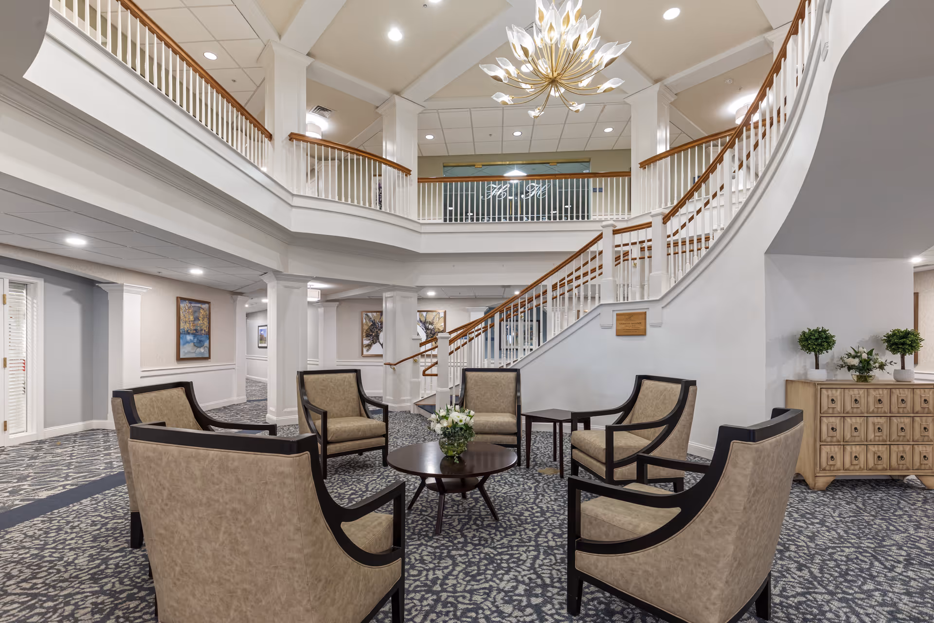 A spacious and elegant common area in a senior living facility featuring a circular arrangement of six beige upholstered chairs around a dark wooden coffee table with a flower arrangement. The area has a patterned carpet, white walls with decorative molding, and a grand staircase with wooden handrails leading to an upper level. A modern chandelier hangs from the ceiling, and there are framed artworks on the walls along with a wooden cabinet with plants on top.