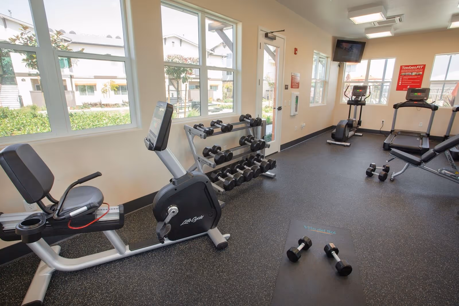 A bright fitness room with large windows showing an outdoor view. The room contains exercise equipment including a recumbent stationary bike, dumbbells on a rack, treadmills, and an exercise bench. A black exercise mat on the floor has the text 'Villa del Sol'.