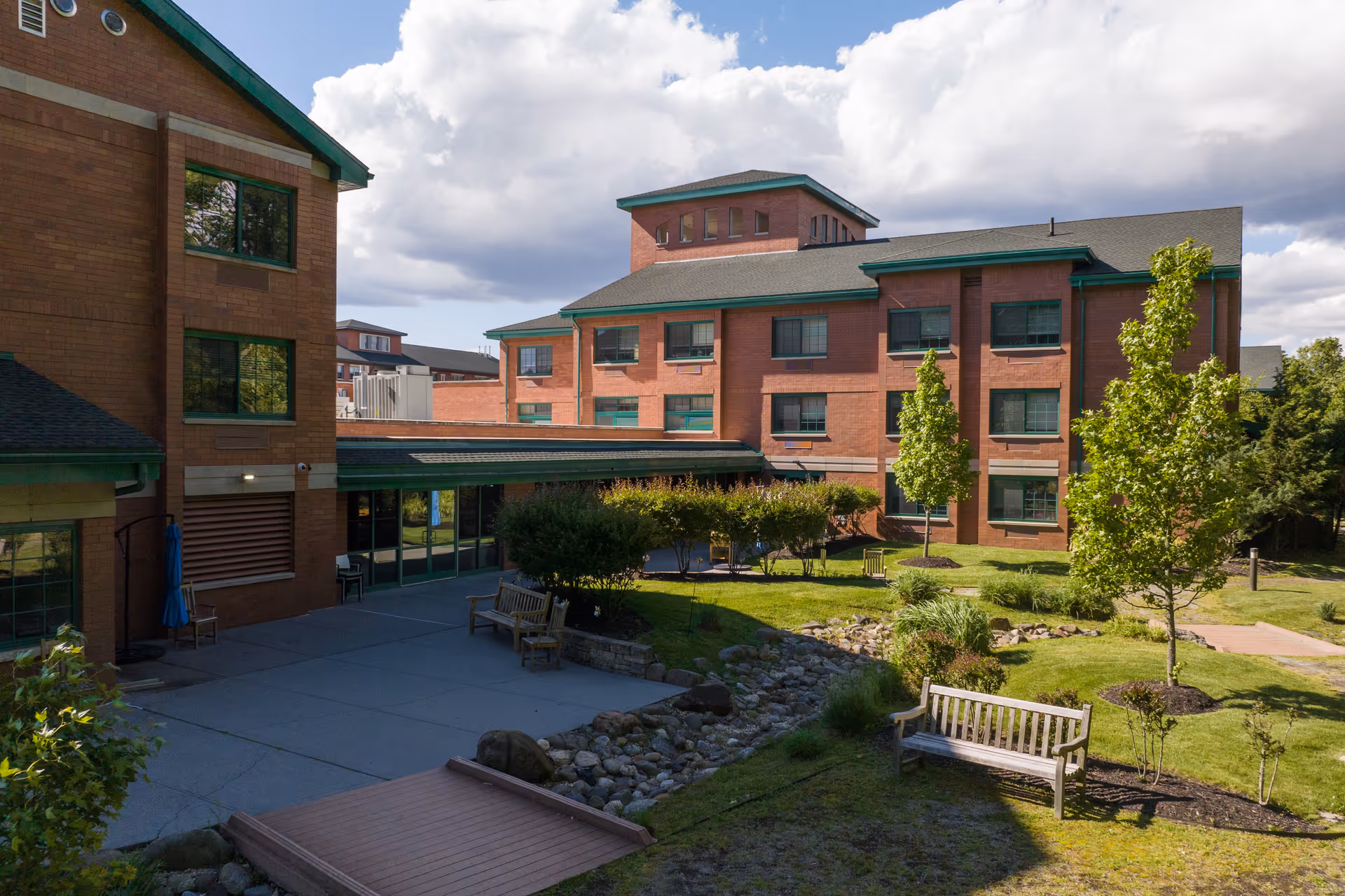 Outdoor view of The Brielle senior living facility showing a brick building with green window frames and a landscaped garden area with trees, benches, a small wooden bridge, and a rock-lined dry creek bed under a partly cloudy sky.